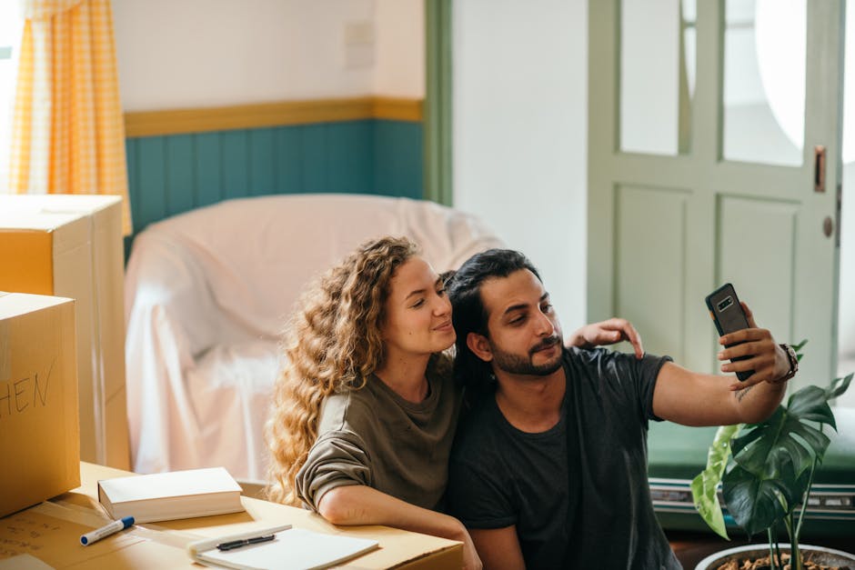 Happy young couple sitting together on floor and taking selfie on smartphone while preparing for relocation in modern apartment