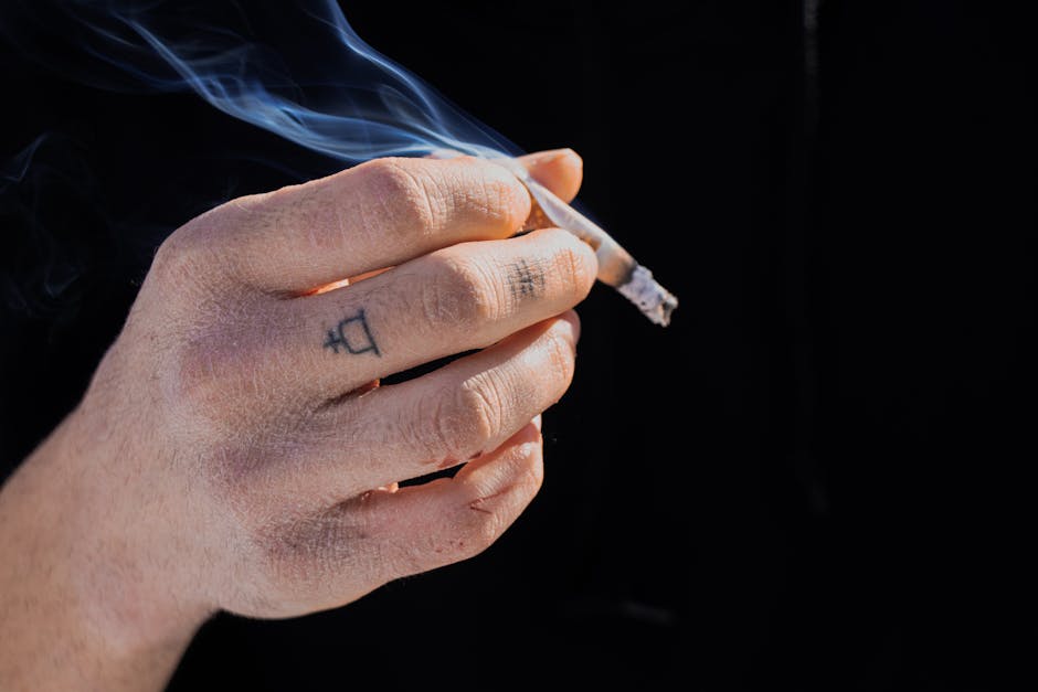A detailed view of a tattooed hand holding a smoking cigarette, emphasizing the act of smoking.