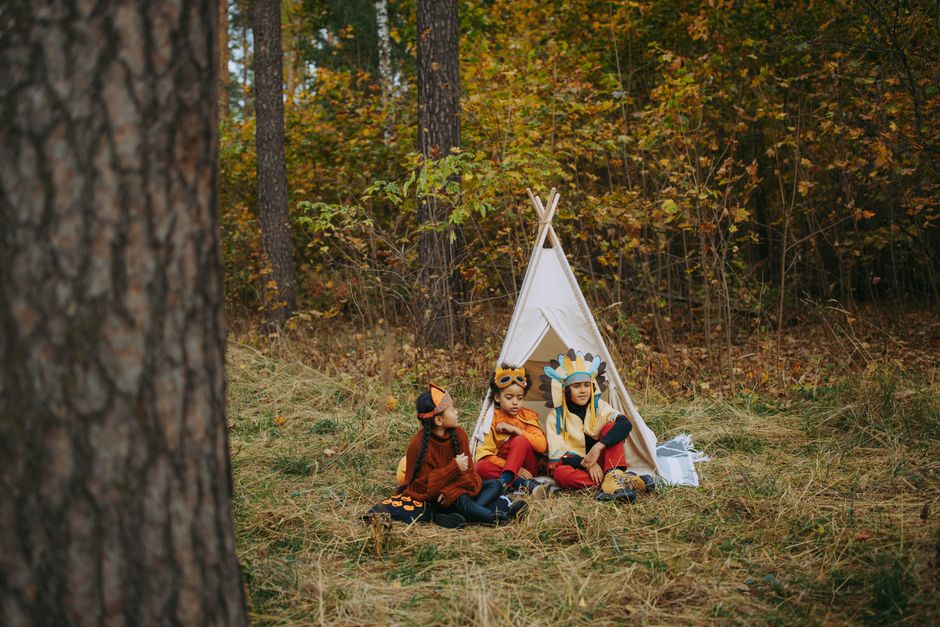Three children in costumes sitting by a tent in an autumn forest, enjoying outdoor playtime.