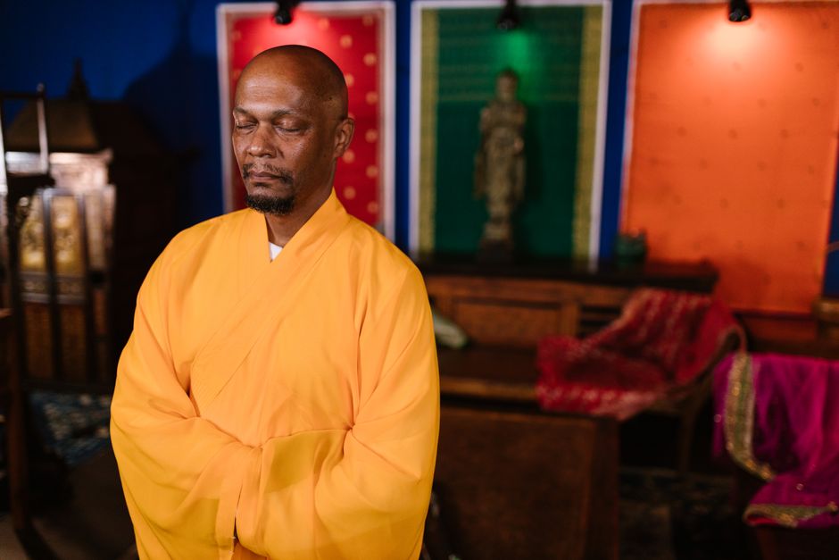 A peaceful Buddhist monk in yellow robe meditating indoors, eyes closed, with colorful backdrop.