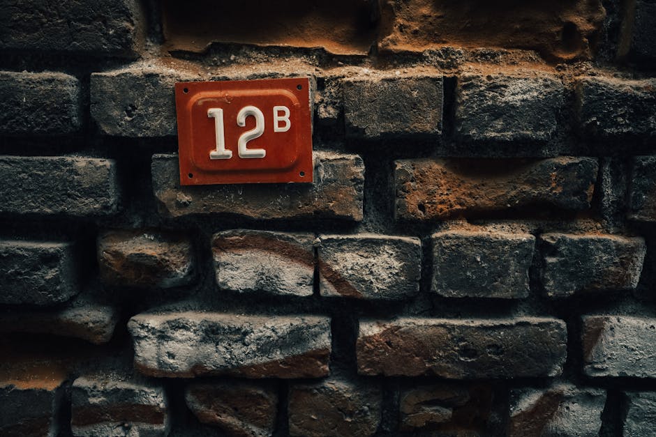 Close-up of a vintage red number plate '12B' mounted on a rustic brick wall.