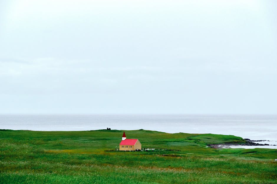 A serene landscape featuring a solitary church with a red roof by the sea, surrounded by lush greenery.