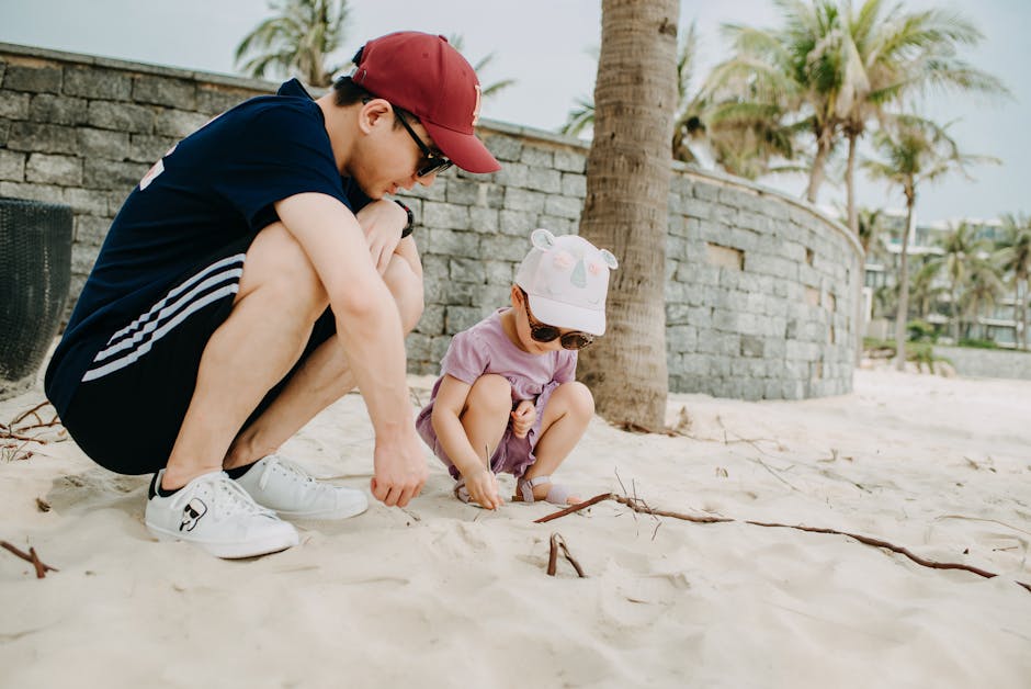 Full body of positive young Asian man wearing red cap playing with child on sandy shore with palms on background on clear day