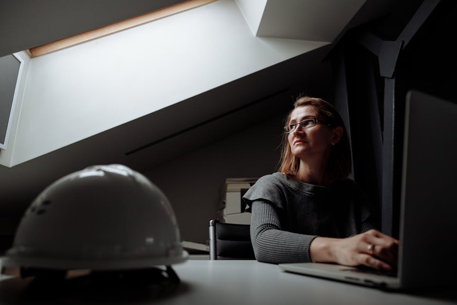 Middle-aged woman in a modern office, thoughtful at work with laptop and hard hat.