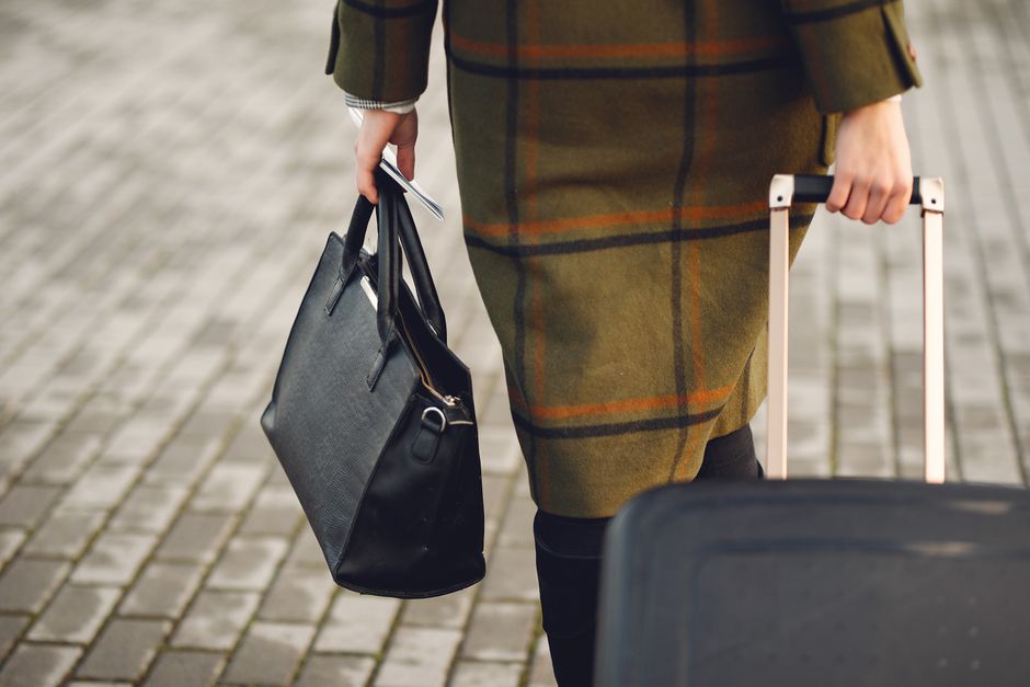 Back view of unrecognizable young female traveler in warm trendy plaid coat strolling on pavement with suitcase and bag while taking passport and tickets on city street in cold season