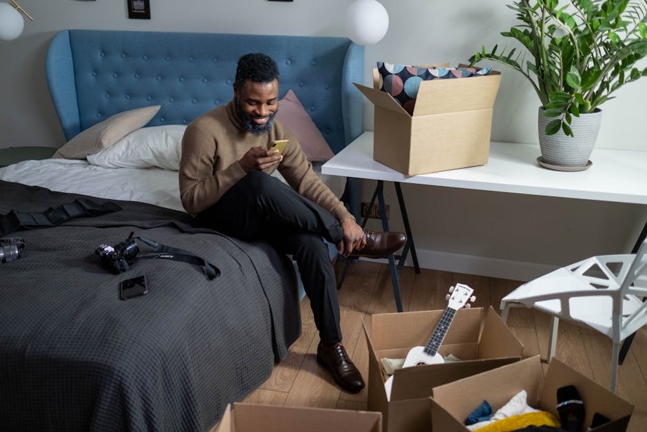Adult man happily texting on smartphone while unpacking in well-decorated bedroom.