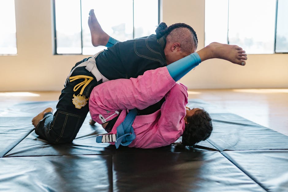 Two athletes engaged in Brazilian Jiu-Jitsu grappling on mats inside a sunlit gym.