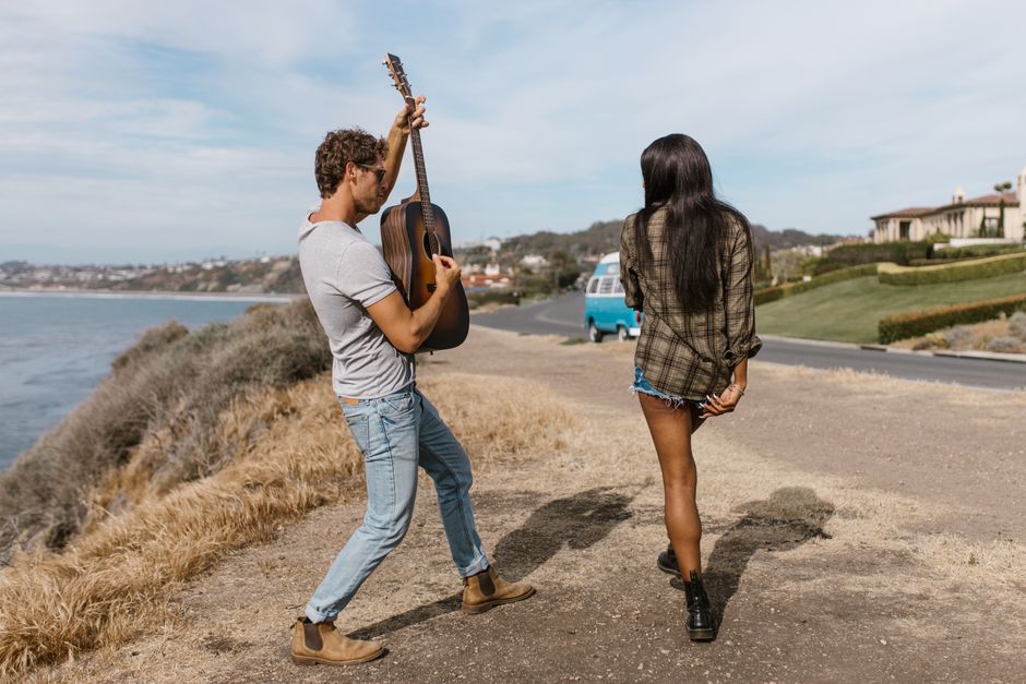 A relaxed young couple on a coastal road trip, with the man playing guitar and a VW van in the background.