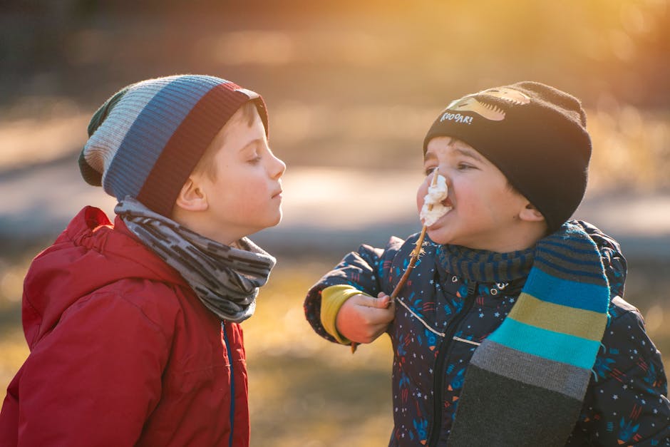 Two young boys wearing winter gear enjoying roasted marshmallows outdoors on a sunny day.