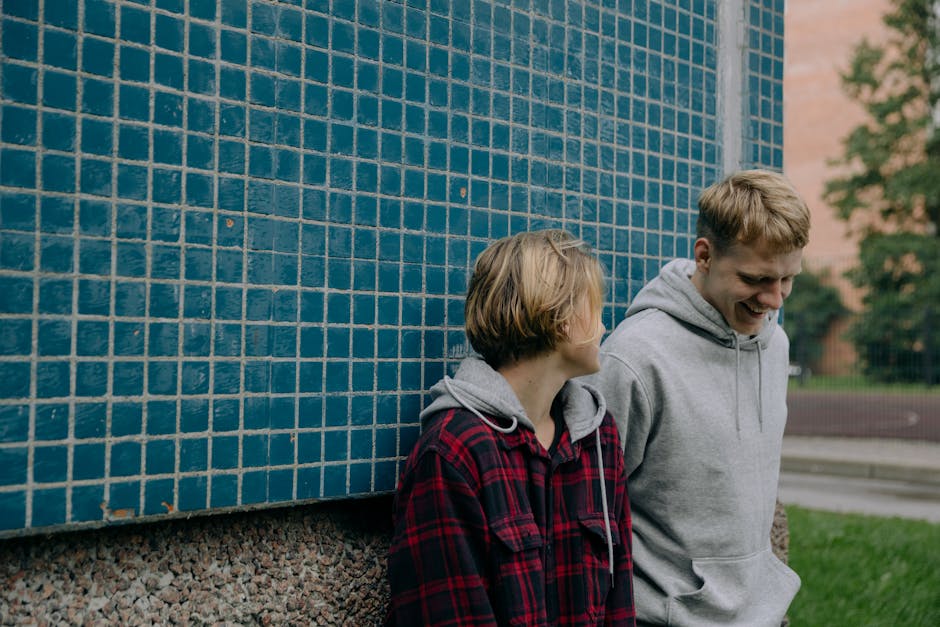 Two young friends in casual attire laughing against a blue tiled wall outdoors, enjoying a casual moment together.