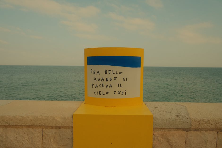 A vibrant yellow post with Italian text by a calm seaside under a clear sky.